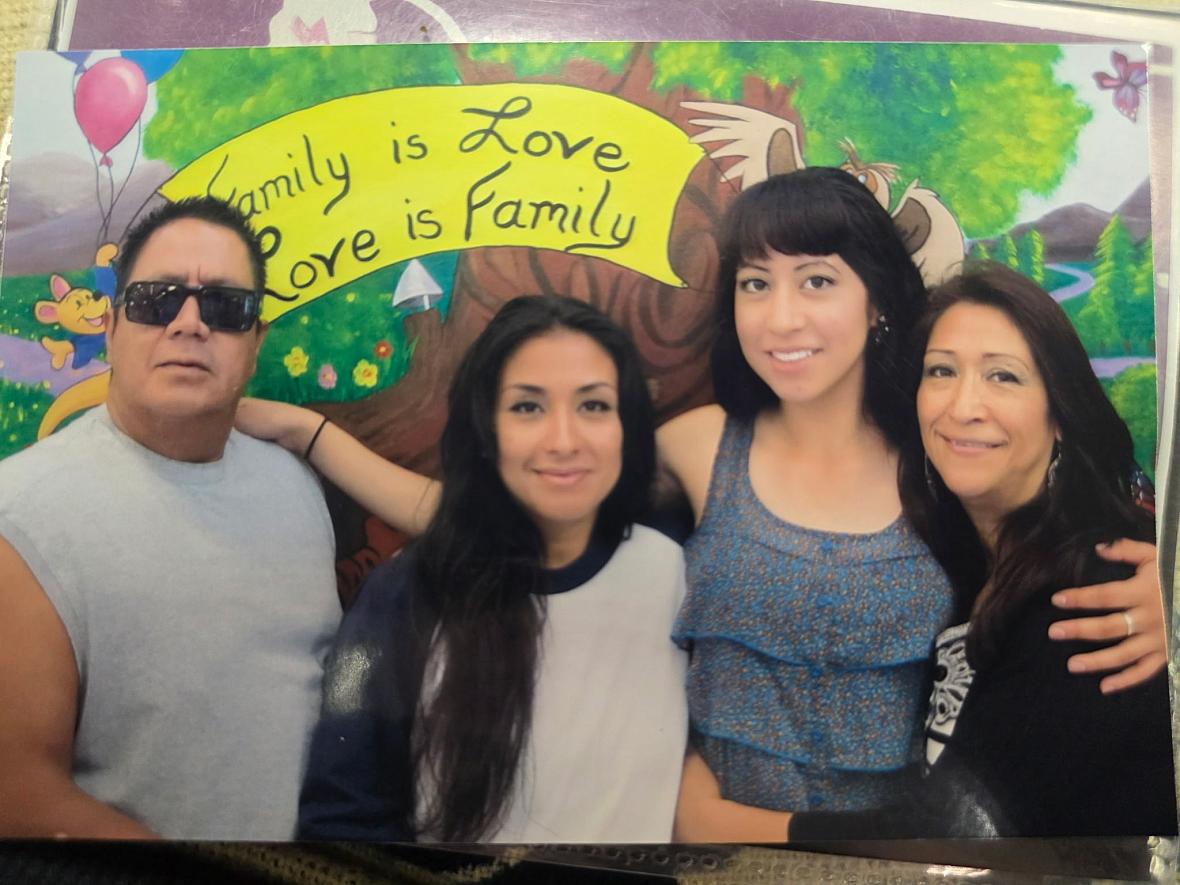 A Latino family of four smiles standing in front of a sign that reads, 'Family is Love, Love is Family'