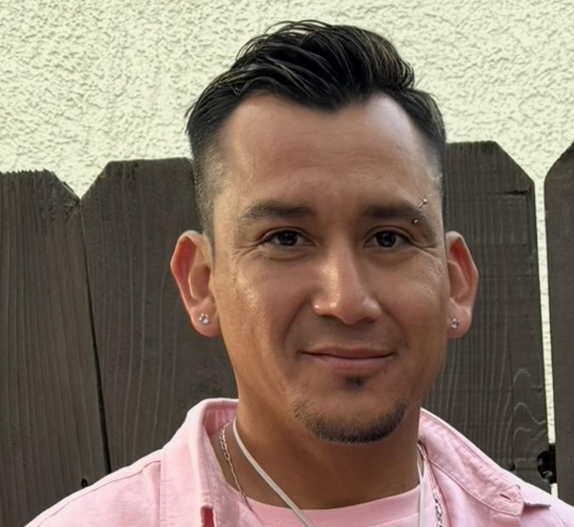 A young El Salvadoran man in a pink shirt stands in front of a brown picket fence