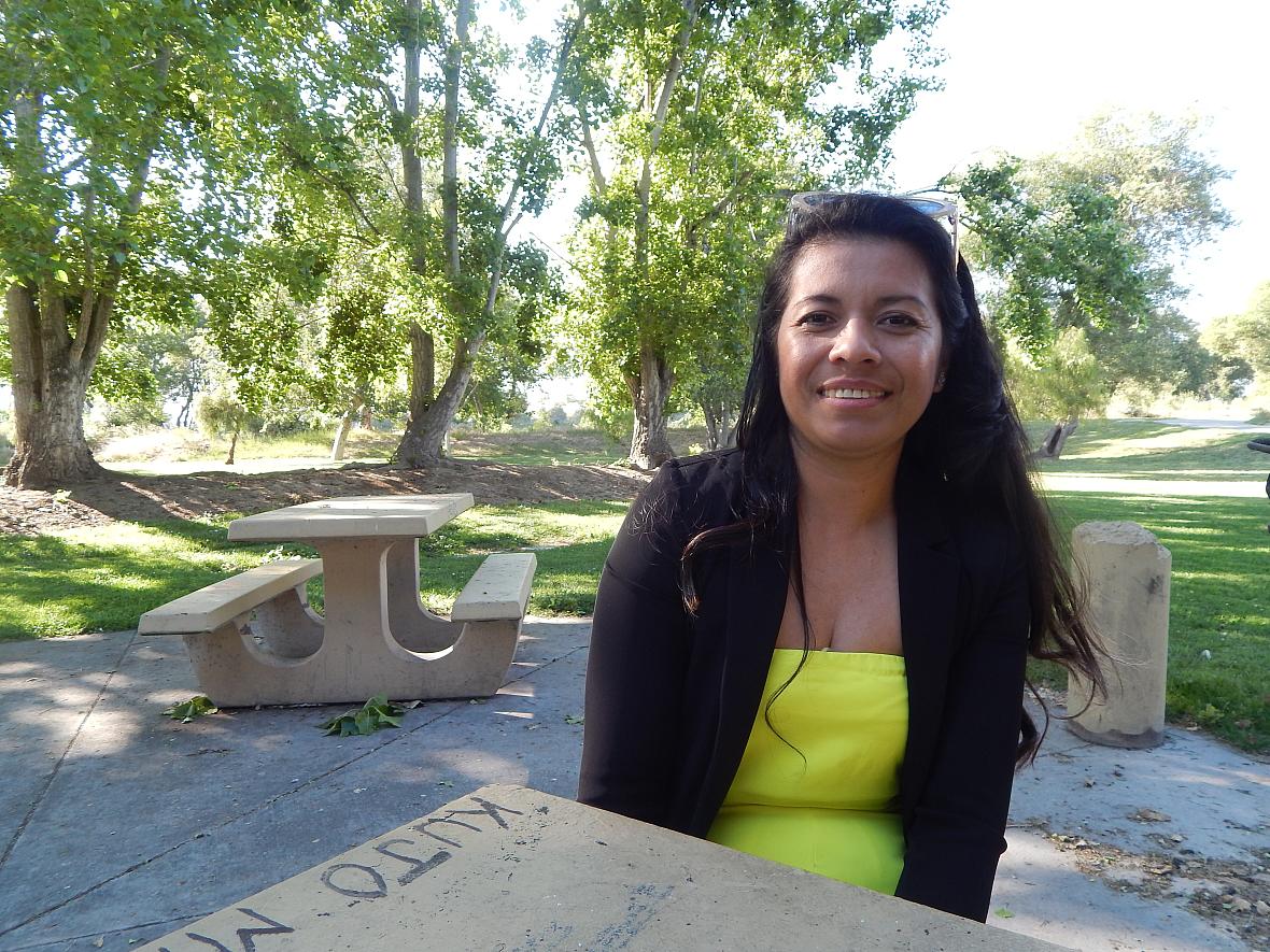 A young woman with long dark hair sits at a picnic bench smiling in a park