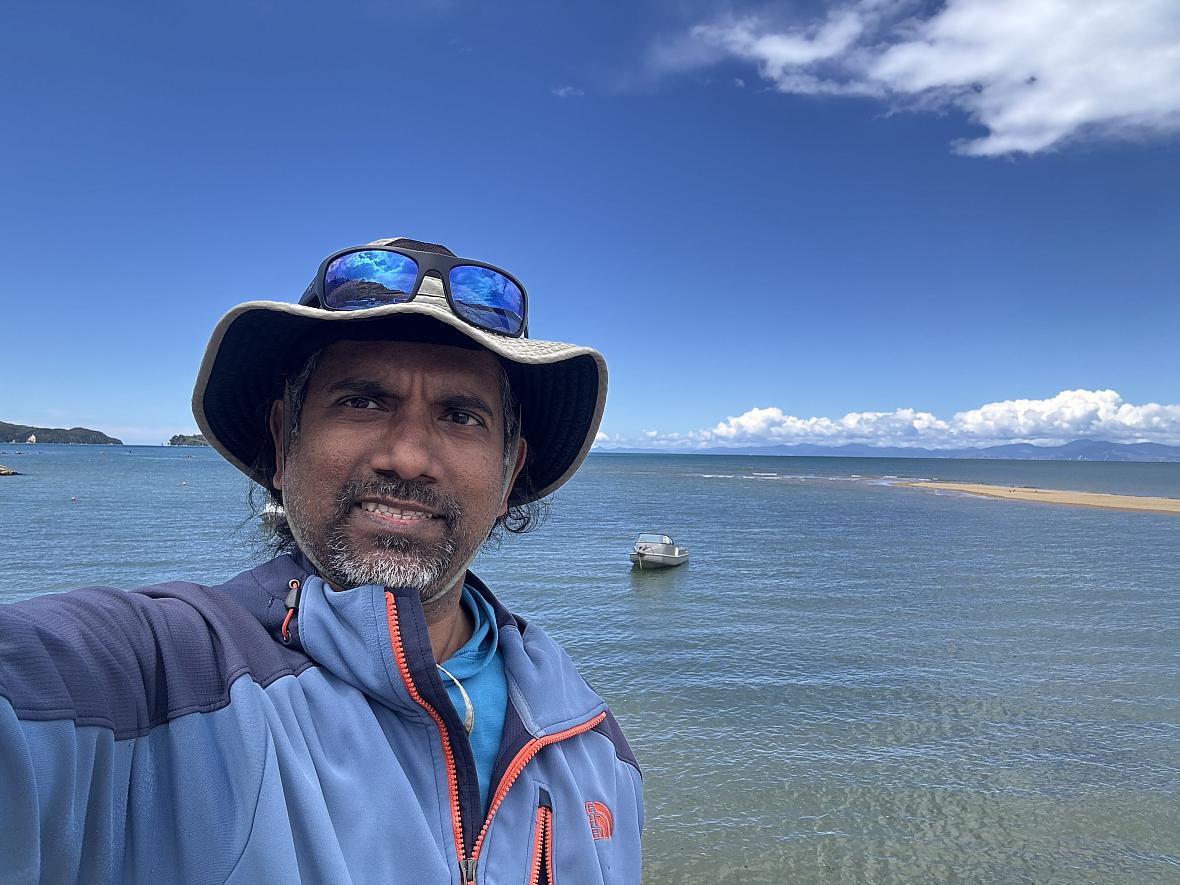 An Sri Lankan Tamil man stands before an expanse of water smiling in a sunhat