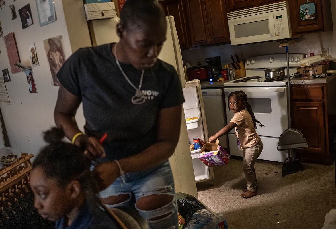 Person doing a child's hair while another child pulls a box out of the refrigerator