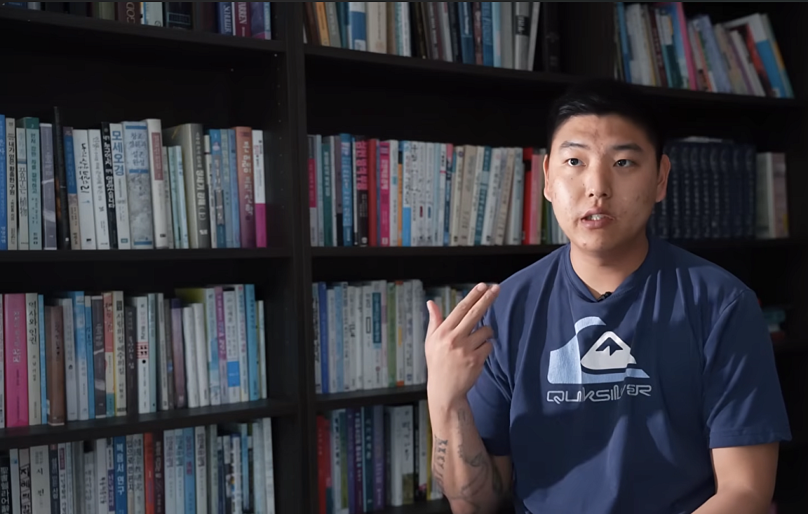 A young Korean man sits in a library making a gun gesture with his hand