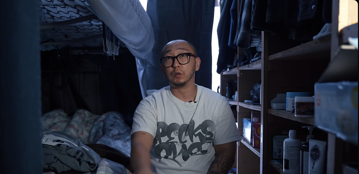 A young Korean man with black glasses sits on his bed