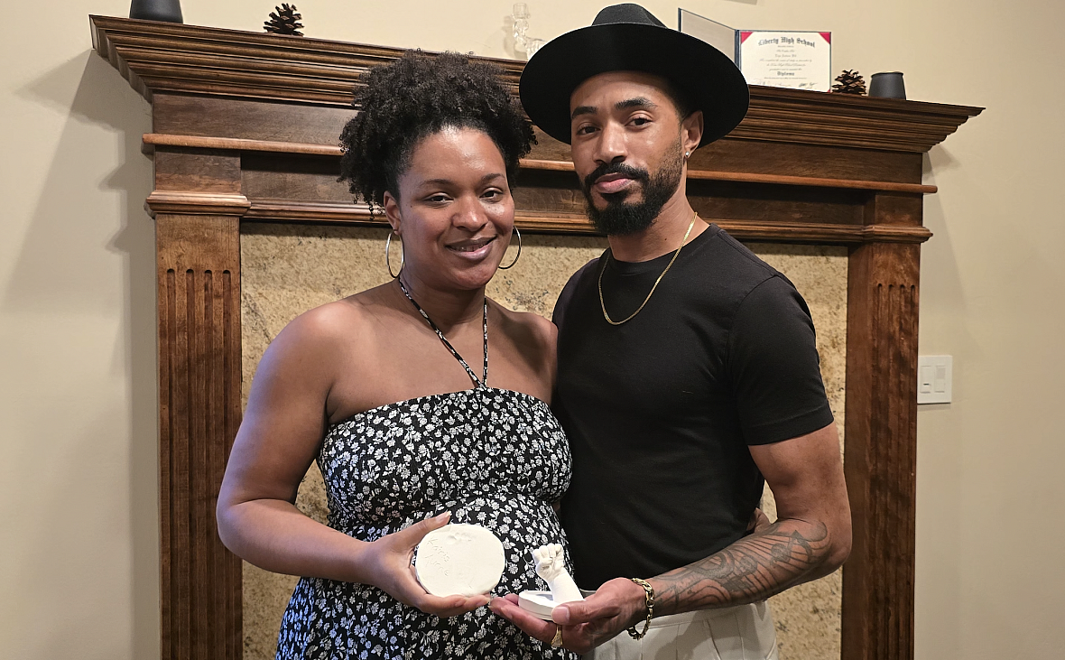 A Black couple stands in front of a wooden mantle holding plaster molds of their late daughters hand and foot.