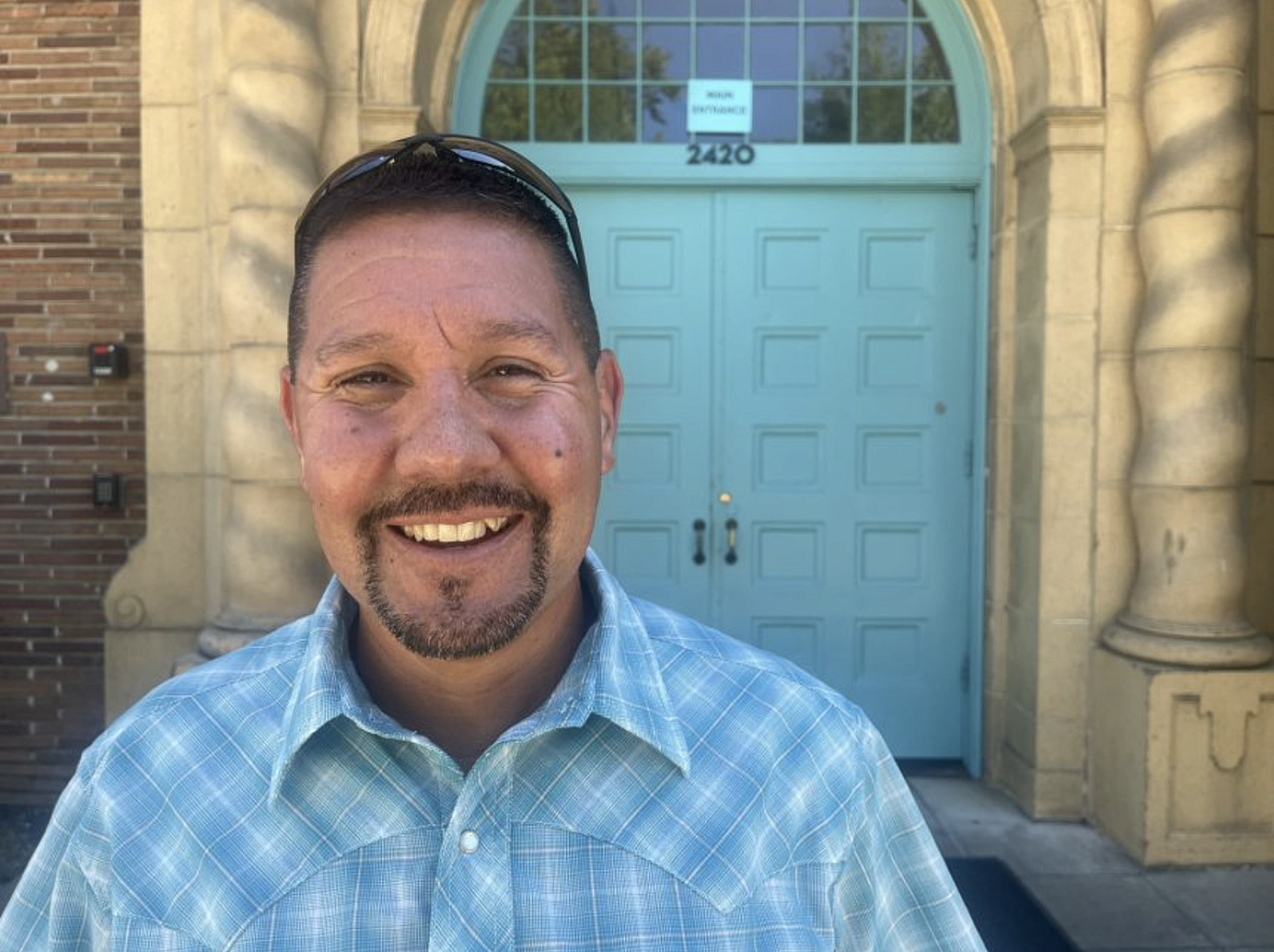 J Vasquez stands in a blue shirt smiling in front of blue double doors.