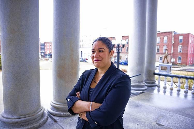 A woman stands confidently with arms crossed on a stone building’s porch, framed by large columns and historic brick buildings in the background.