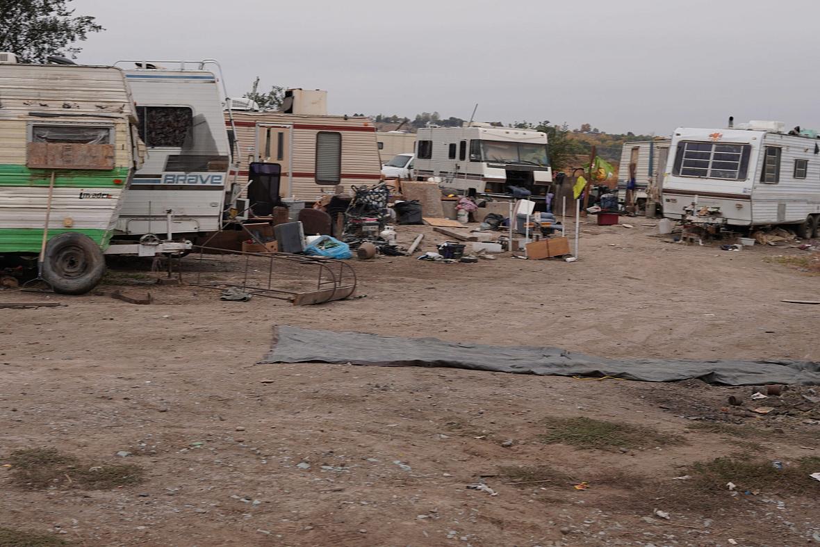 A makeshift encampment of old RVs and trailers surrounded by scattered trash and debris on a dirt lot, indicating homelessness and poverty.