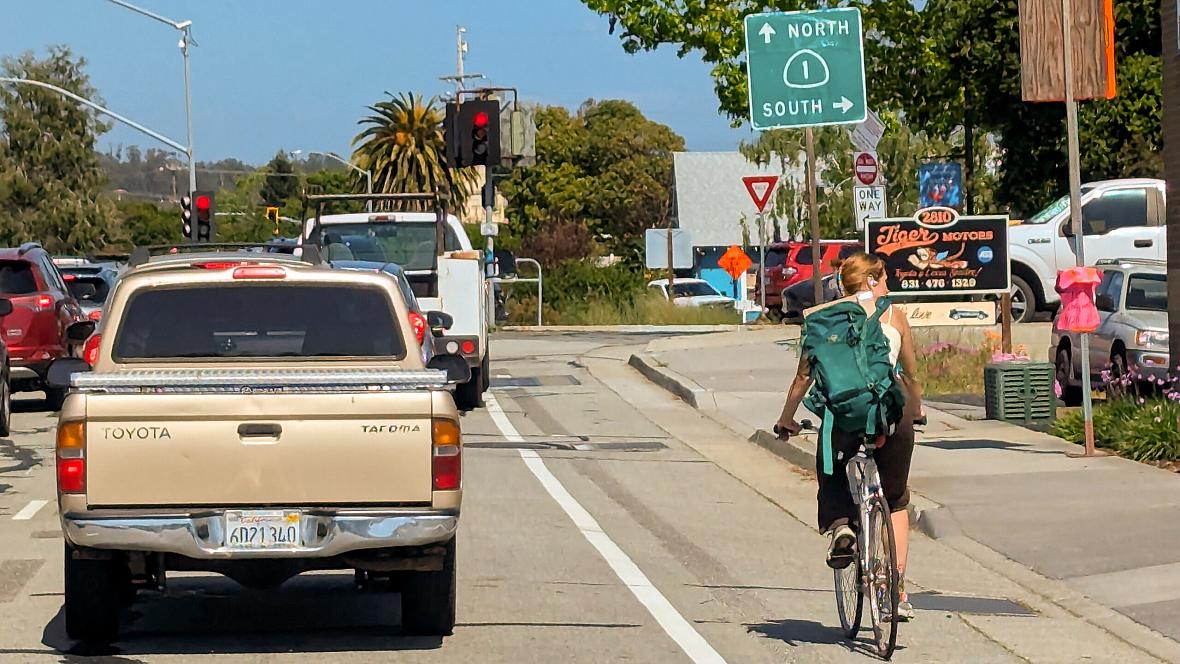 A woman wearing a backpack rides a bicycle in a designated bike lane beside a beige pickup truck at a traffic light intersection. Overhead signs indicate directions for Highway 1 North and South. Traffic is visible in multiple lanes, and there are businesses, including “Tiger Motors,” on the right side of the road. The weather is sunny with clear skies.