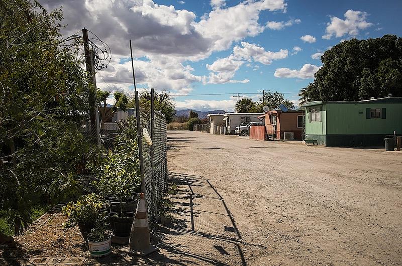 Image of dirt road on a sunny day with houses on its right and trees on left.