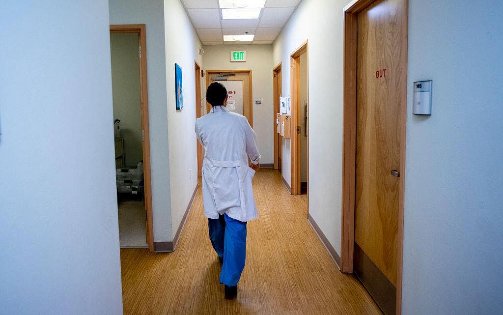 A medical professional in a white coat walks down a brightly lit hallway in a clinic or hospital, with doors labeled “OUT” and an exit sign visible ahead.