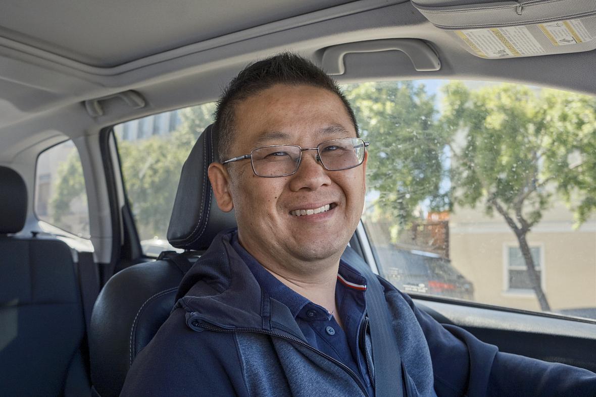 Person sitting in the driving seat of a car with a blue jacket and well cropped hair with spectacles