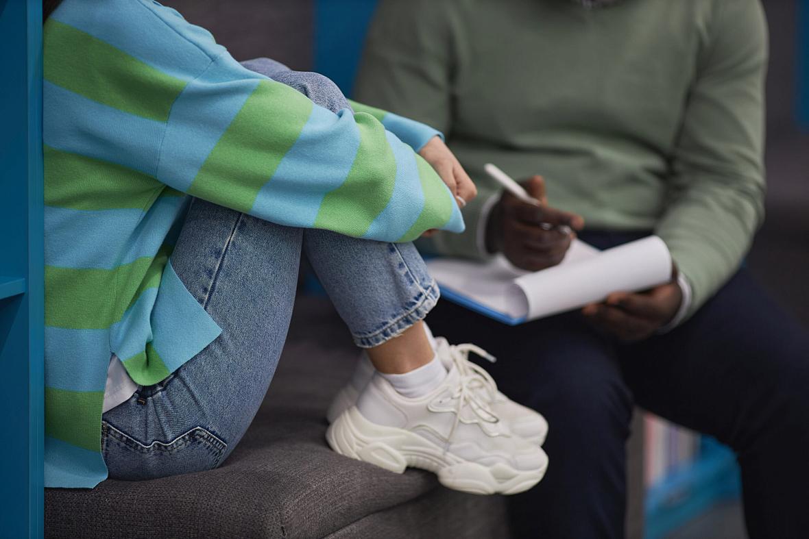 Person sitting with legs close to body, wearing white shoes along with a person writing notes in the background