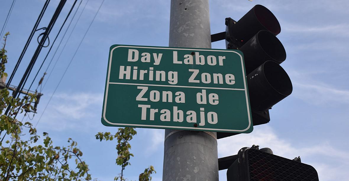 A bilingual street sign reads “Day Labor Hiring Zone / Zona de Trabajo” near a traffic light.