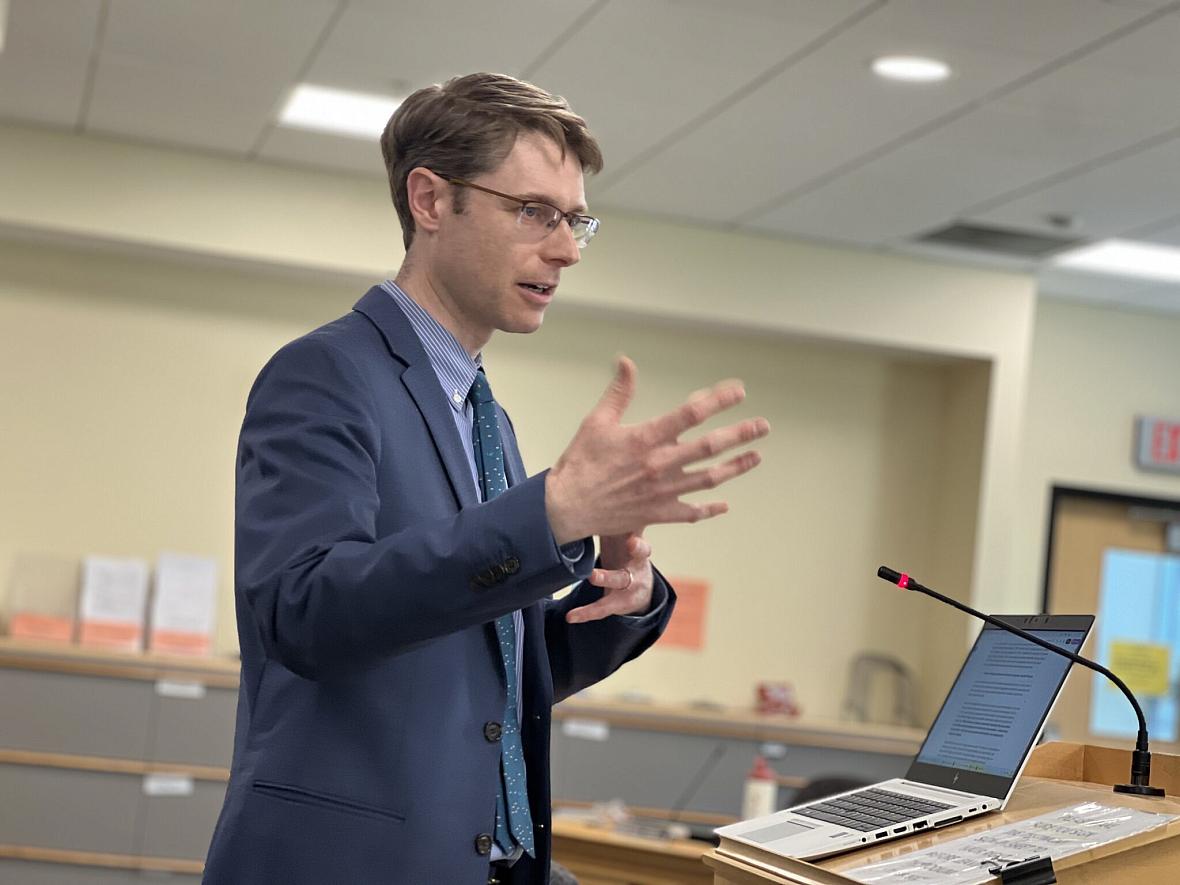 A man in a suit speaks at a podium with a laptop and microphone in a meeting room.
