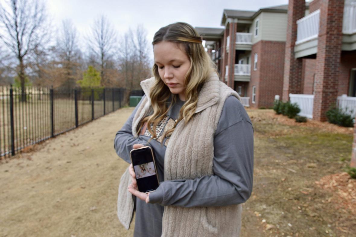 A woman outdoors holds a phone displaying a photo and looks down at it, with apartments and leafless trees in the background.