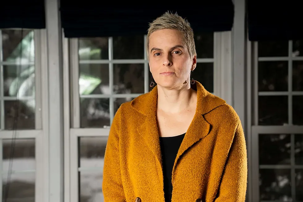 Close-up of a woman in a mustard coat standing indoors with a serious expression, in front of large windows.