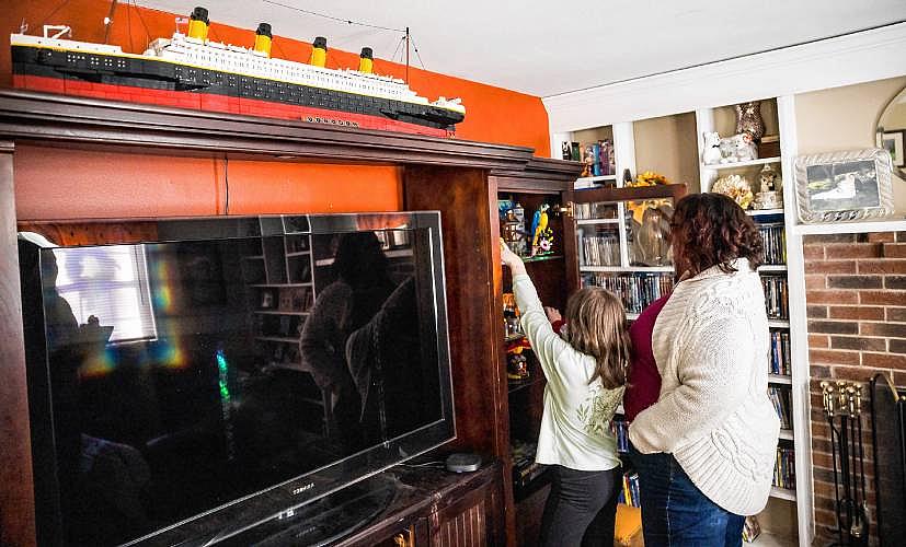 A woman and a child stand in a living room reaching for toys on a shelf beside a TV cabinet, with a large model ship displayed above.