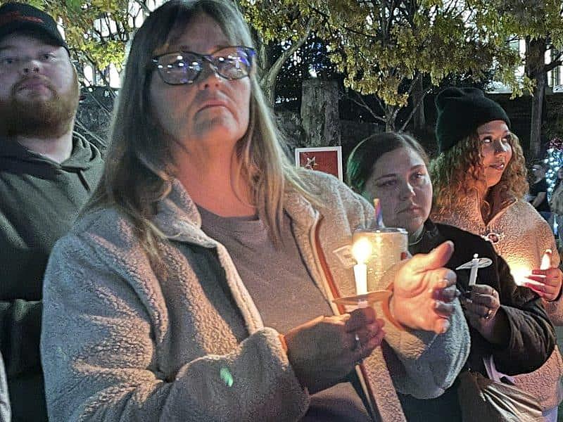 A group of people holds candles at a nighttime vigil; a woman in the foreground wears glasses and a fleece jacket, looking solemnly ahead.