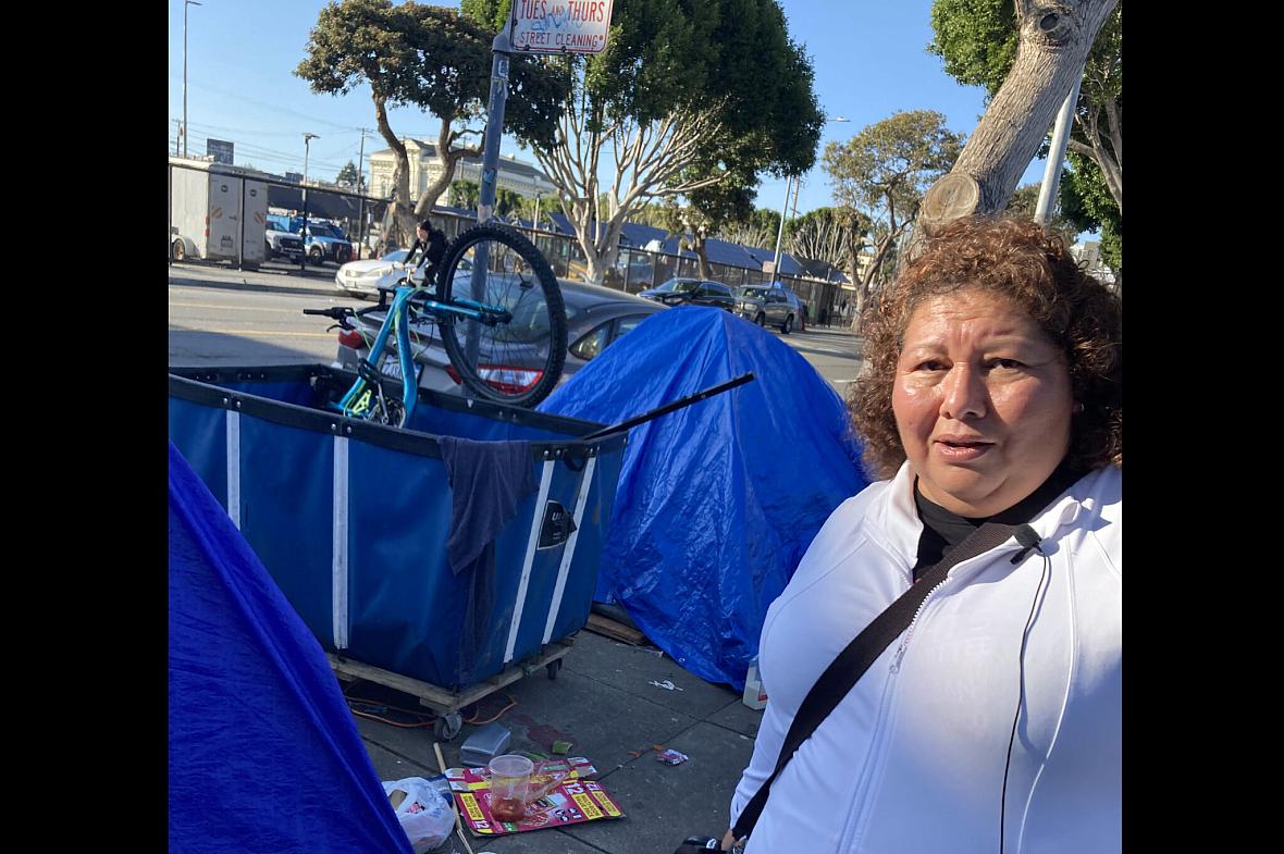 Person with microphone standing in front of blue tent like tarp on the sidewalk