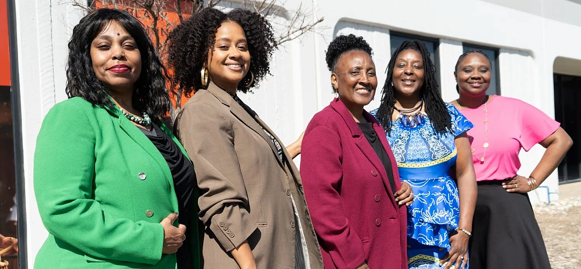 Five women standing outside, smiling in colorful professional outfits.