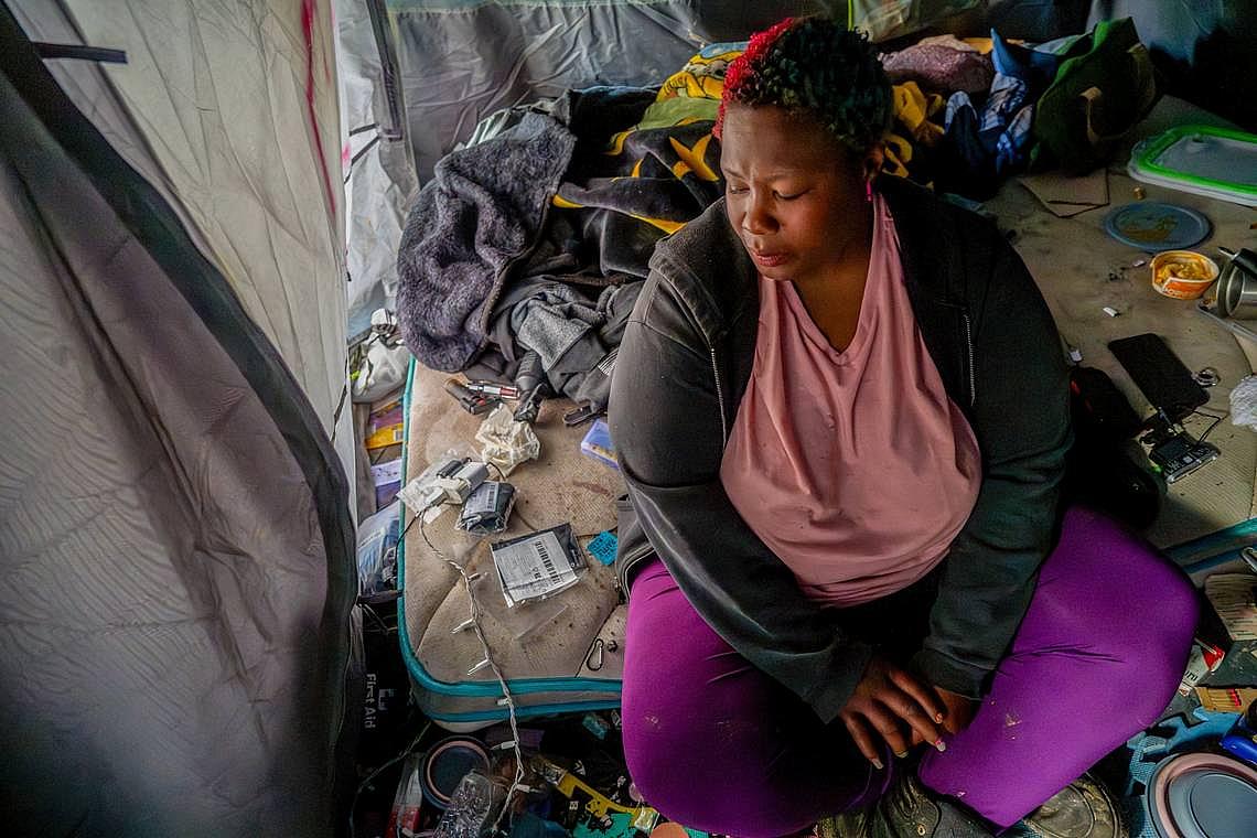 Woman sits inside a cluttered tent, looking down, surrounded by personal belongings.