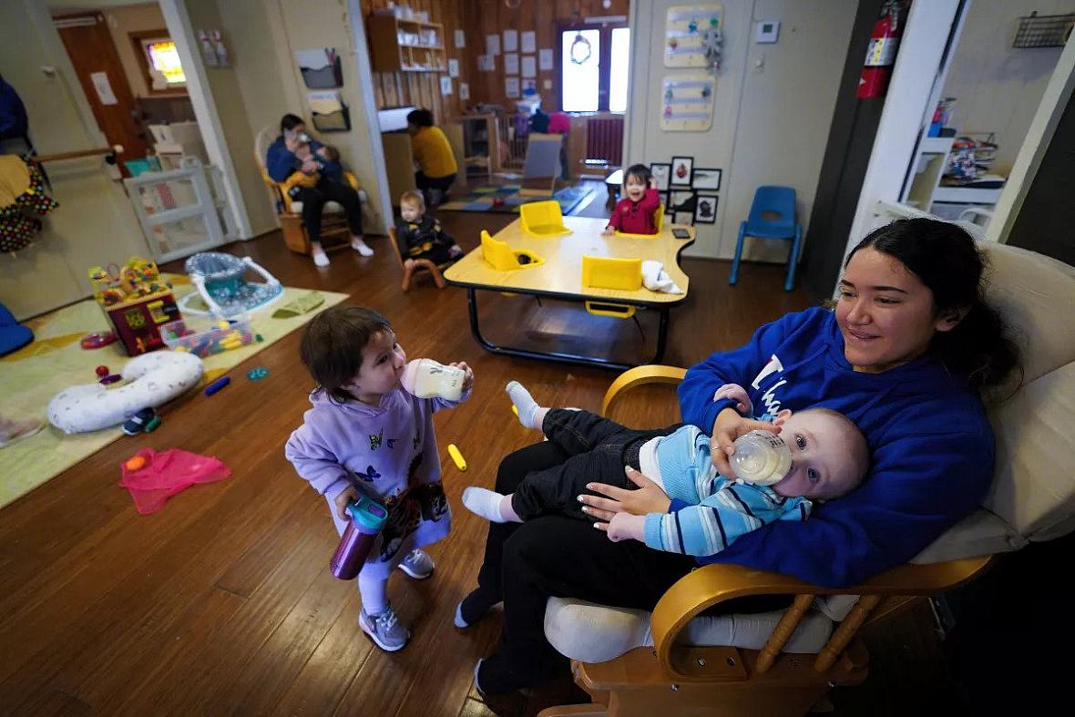 A girl feeding a baby and another child in front of her drinking milk from sippy cup