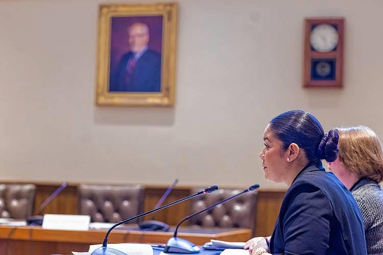 Person in courtroom, sitting at a desk and speaking on a microphone
