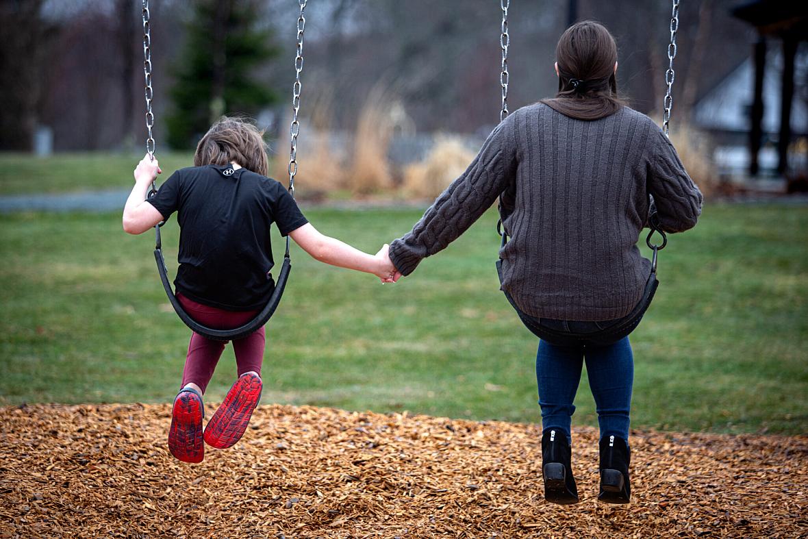 A child and an adult sit on swings holding hands, seen from behind, in a grassy playground with wood chips on the ground.