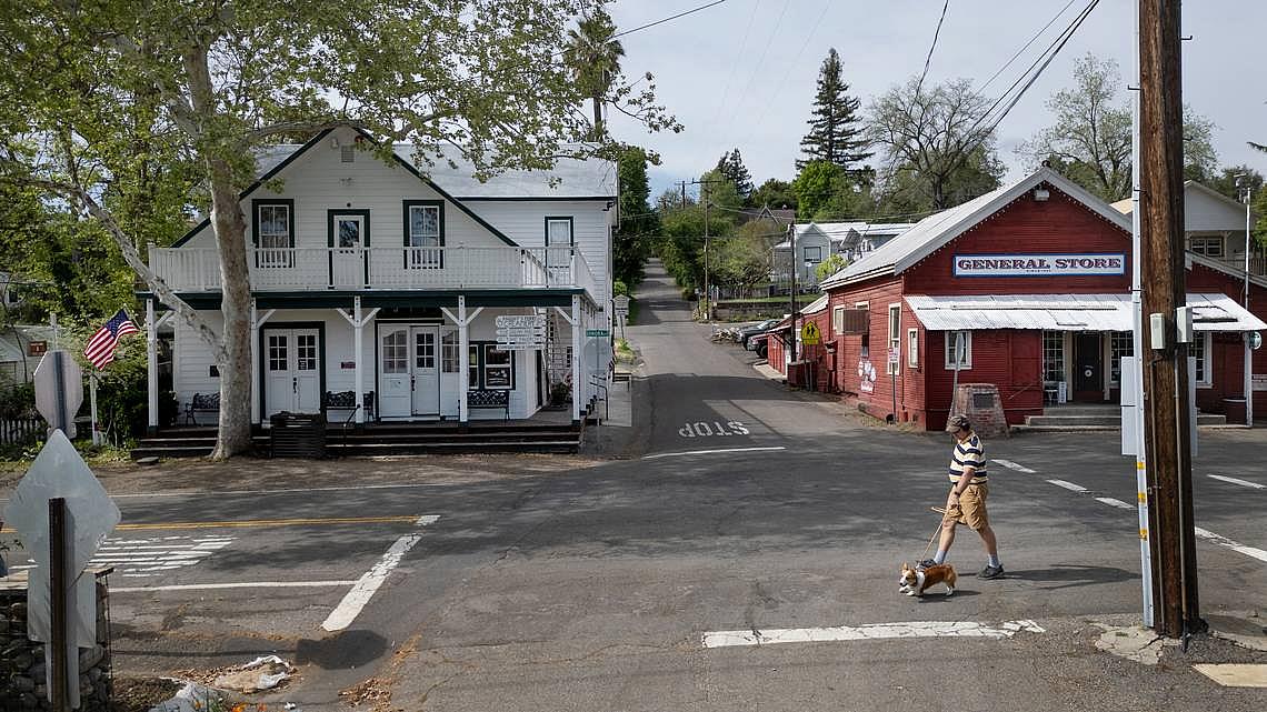 Man walking a dog past a general store in a quiet small town.
