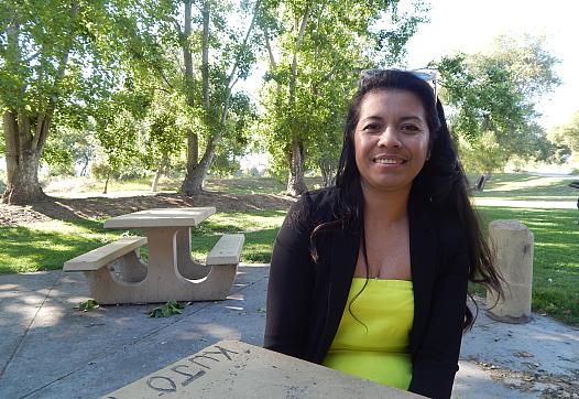 A young woman with long dark hair sits at a picnic bench smiling in a park