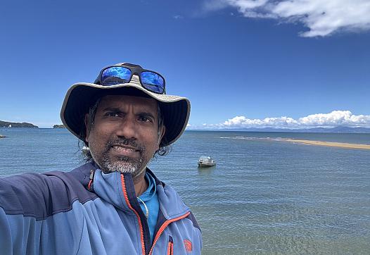 An Sri Lanken Tamil man stands before an expanse of water smiling in a sunhat