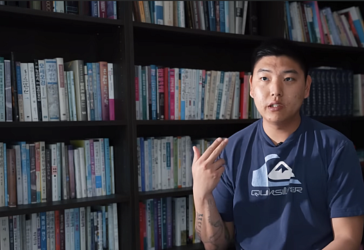 A young Korean man sits in a library making a gun gesture with his hand