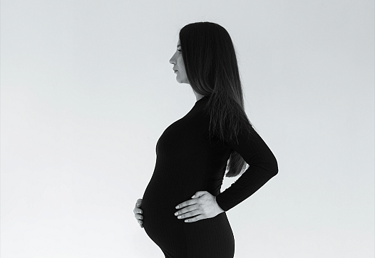 A young Asian Indian woman with long dark hair stands in profile, pregnant, in a tight black dress