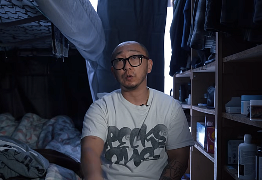 A young Korean man with black glasses sits on his bed