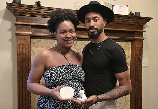 A Black couple stands in front of a wooden mantle holding plaster molds of their late daughters hand and foot.