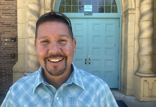 J Vasquez stands in a blue shirt smiling in front of blue double doors.