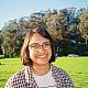 A young women with a brown bob and glasses stands smiling in a field and a plaid shirt.