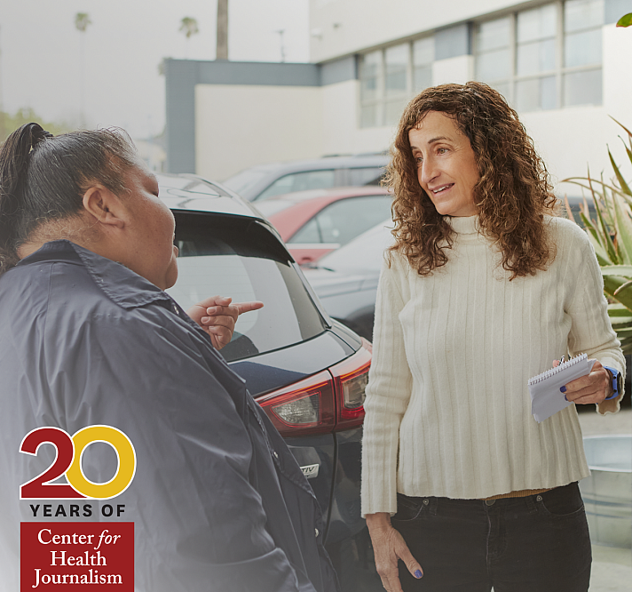 A woman with curly brown hair, wearing a white ribbed sweater, holds a notepad and pen while speaking with another person outside in a parking lot. The second person, dressed in a dark jacket, gestures with their hand while facing the woman. Cars are visible in the background, and the conversation appears informal and engaged. In the bottom-left corner, a logo reads "20 Years of Center for Health Journalism" in red and gold.