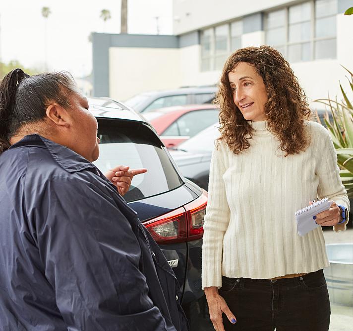 A person with curly hair holding a notepad speaks with a woman in a parking lot, listening as she gestures during their conversation.