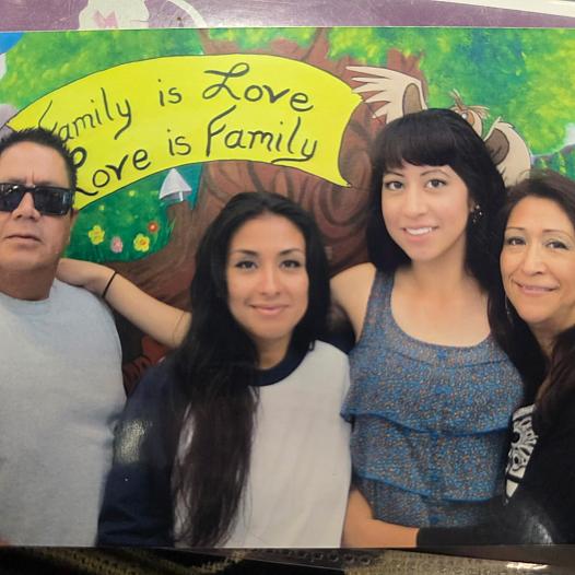 A Latino family of four smiles standing in front of a sign that reads, 'Family is Love, Love is Family'