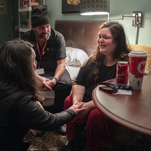 A woman smiles while speaking with two others in a cozy room. One person holds her hands supportively. A soda, coffee cup, and keys rest on the nearby table.