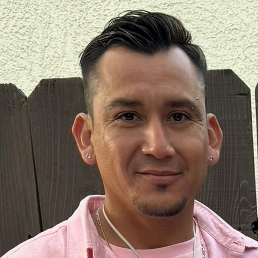A young El Salvadoran man in a pink shirt stands in front of a brown picket fence