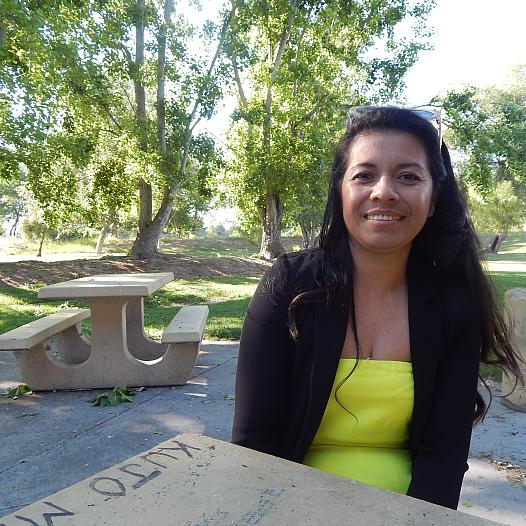 A young woman with long dark hair sits at a picnic bench smiling in a park