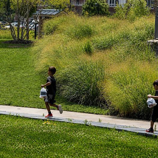 Three children run through a park with bags in their hands.