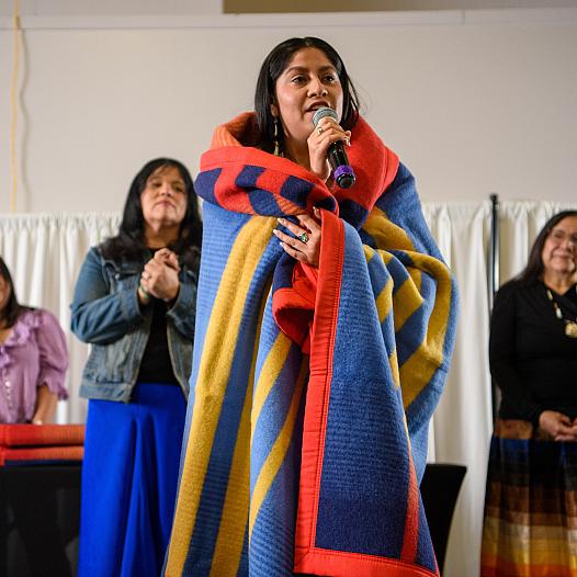 Huwighurruk Tribal Health Program graduate Desirae Barragan, wrapped in a colorful blanket, introduces herself in her native language during the Huwighurruk blanket ceremony in 2025. 