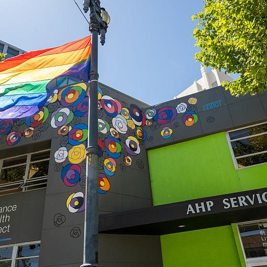 A rainbow flag flies in front of two buildings in San Francisco, including UCSF Alliance Health Project