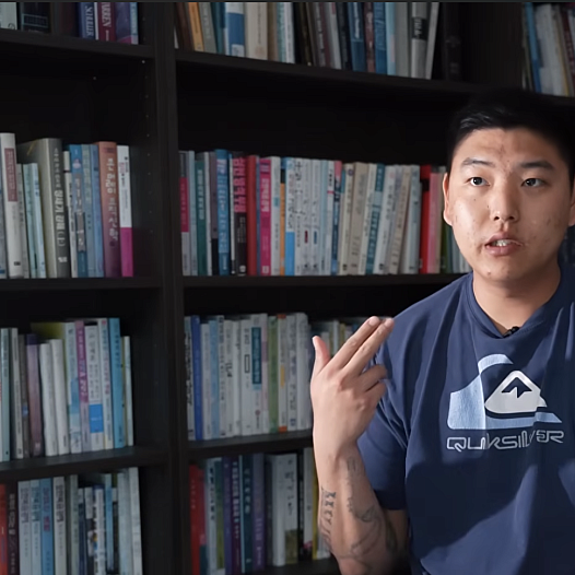 A young Korean man sits in a library making a gun gesture with his hand