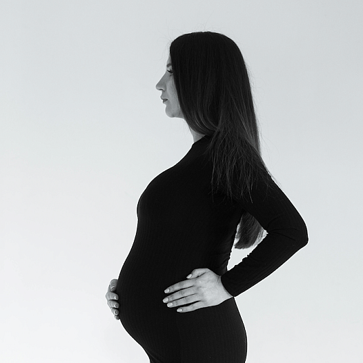 A young Asian Indian woman with long dark hair stands in profile, pregnant, in a tight black dress