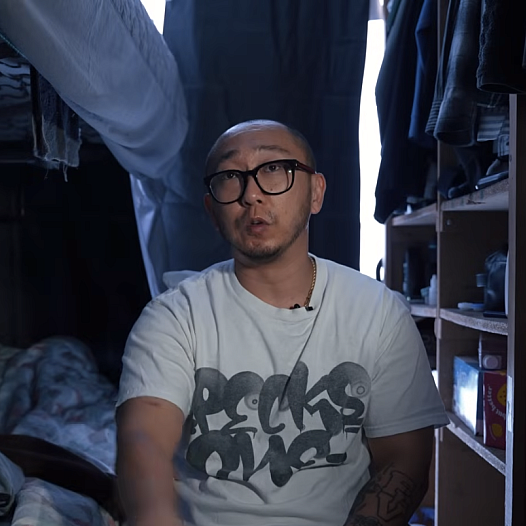 A young Korean man with black glasses sits on his bed