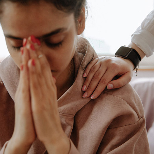 A young South Asian Indian Woman buries her face in her hands as a therapist comforts her.
